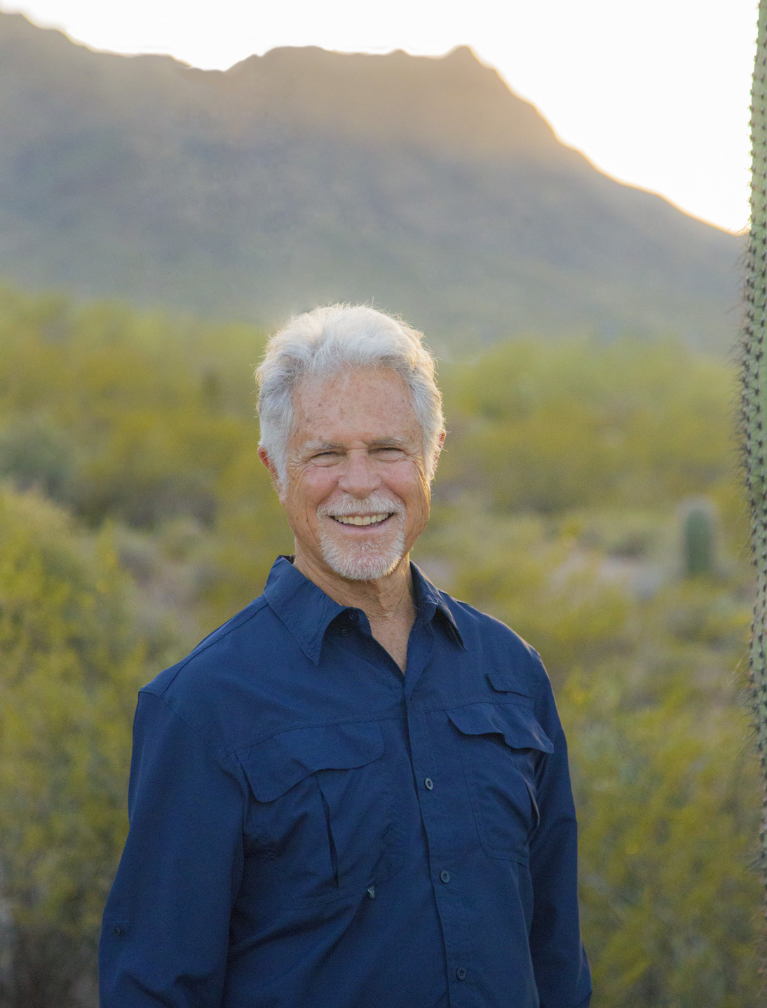 A man with white hair and blue shirt standing in front of trees.